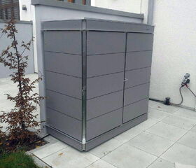 . The image shows a gray metal utility box or storage cabinet placed outside, next to a wall. It's located on a tiled patio area. There is a small plant nearby and a hose attached to a nearby tap on the wall. The setting appears to be part of a residential or commercial outdoor space.