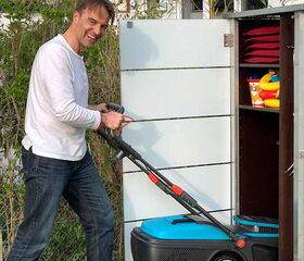 . The image shows a man smiling and pushing a lawn mower. He is standing on grass next to a storage shed. The shed has shelves with some items, including what appear to be red cushions and colorful toys. The setting is outdoors, adjacent to a building.