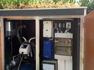 . The image shows an equipment cabinet or enclosure, likely for a pool or spa. It contains various machinery and devices, such as pumps, filters, control panels, and wiring. The setup is organized within a metal frame, with components neatly arranged. The enclosure also has a green roof with plants.