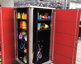 . The image shows an open red storage cabinet containing various toys and outdoor play equipment. Inside, there are:

1. Watering cans and buckets.
2. Small bicycle or tricycle.
3. Assorted balls, including whiffle balls.
4. Stacked containers or tubs.
5. Play toys and equipment.

The cabinet is standing on what appears to be a patio or outdoor area with a protective roof structure above.