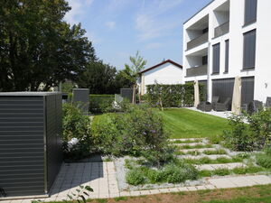 . The image depicts a residential outdoor area featuring:

1. A modern white building with balconies.
2. A manicured green lawn.
3. Small garden plots with plants.
4. Outdoor furniture under covered patio areas.
5. Two gray garden sheds.
6. Paved stone pathways.
7. Trees and hedges providing natural greenery.

The setting appears to be calm and well-maintained.