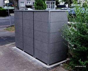 . Drei graue Mülltonnenboxen auf einem Gehweg neben grünen Büschen in einer städtischen Umgebung.