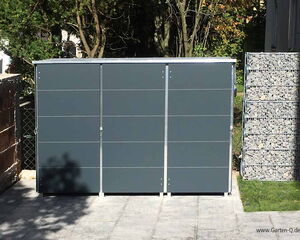 . The image shows a modern storage unit or garden shed with a sleek design, situated outdoors. It has a dark gray color with a metal frame, and it's positioned on a paved surface. To the right, there is a structure filled with stones, likely for decorative or structural purposes, and trees are visible in the background.