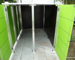 The image shows a storage unit with open doors, revealing a spacious interior. The exterior is green with horizontal lines, and the frame appears to be made of metal. The floor is paved, and there is vegetation visible in the background on the right. The structure seems suitable for storing outdoor items.
