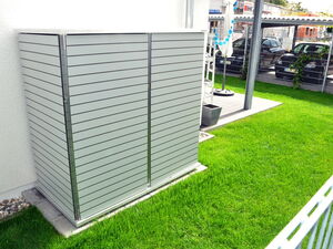. The image shows an outdoor area with a well-maintained grass lawn. In the foreground, there is a green storage box or shed with horizontal slats, likely used to cover or store materials outside. In the background, there are cars parked behind a wire fence, and some outdoor furniture, including an umbrella. The overall setting appears to be a residential or commercial area with a neat and organized appearance.