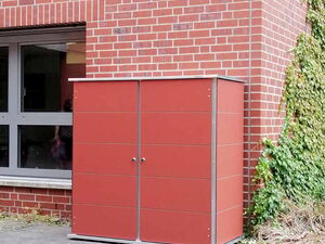 . The image shows a brick wall with a red metal outdoor storage cabinet placed against it. The cabinet has two doors and is on a concrete patio area with some overgrown plants nearby. There's also a window to the left of the cabinet.