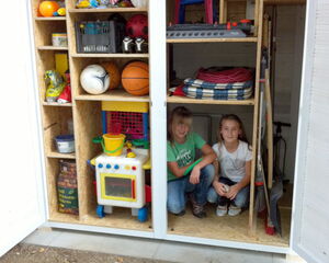 The image shows an open wardrobe or storage unit with multiple shelves containing various items. On the left side, the shelves hold items like a football, basketball, other sports equipment, toys, a colorful plastic kitchen set, and some household supplies. On the right side, two children are sitting inside, and the shelves above them contain items like folded mats, a garden hose, small chairs, and various tools. The overall scene appears organized, with a mix of toys and household equipment visible in the storage units.