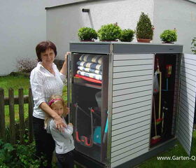 . The image shows a woman and a child standing next to an outdoor storage cabinet. The cabinet is open, displaying various garden tools and supplies inside, such as brooms, rakes, and folded towels. On top of the cabinet, there are several potted plants. The setting appears to be a garden or backyard.