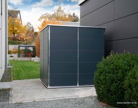 . The image shows a modern garden shed situated on a paved area next to a building. The shed has a sleek, minimalist design with dark panels and a metal frame. It's positioned next to a hedge, and there's a lawn area leading to other garden elements like a pergola or gazebo. The background features autumn foliage.