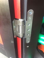 This image shows a close-up of a door hinge. The hinge is metallic and attached to a door, which appears to be made of wood or a similar material. There are multiple screws visible that attach the hinge to the door and frame. The door has a red edge, and part of a green object is visible in the background.
