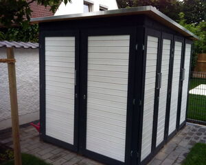 The image shows a small outdoor storage shed. It appears to have multiple doors with white panels and dark frames. The shed is situated on a paved area and is likely used for storing garden tools or equipment. There is a house and some greenery in the background.