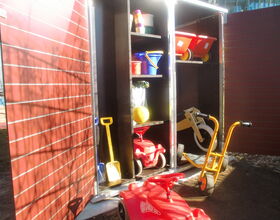 . The image shows an open storage cabinet with various outdoor toys and equipment. Here's a list in array format:

1. Red ride-on toy car
2. Yellow shovel
3. Children's bicycles/trikes
4. Red and yellow wheelbarrow
5. Assorted buckets
6. Balls
7. Toy construction helmets
8. Various containers

The cabinet appears to be in an outdoor area, possibly a playground or backyard.