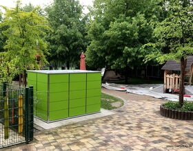 The image shows an outdoor area with several elements:

1. **Green Storage Unit**: A large, green rectangular storage unit or shed.
2. **Playground Equipment**: There appears to be playground equipment, partially visible behind some trees.
3. **Trees**: Several green trees are present, providing shade.
4. **Paved Area**: A paved walkway or patio made of square tiles.
5. **Fence**: A metal fence with colorful poles next to the storage unit.
6. **Ground Cover**: Some areas are covered with a tarp or fabric, likely to protect or mark off sections of the ground.

The setting appears to be a playground or a garden area, possibly in a park or school.