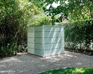 . The image shows a modern, minimalist outdoor storage shed or cabinet situated in a garden. It's surrounded by greenery, including bushes and trees, and is placed on a pebble-covered area. The shed has a sleek, metallic appearance with horizontal lines or slats and appears to be well-integrated into its garden environment.