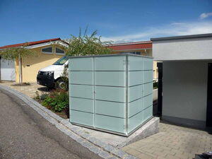 . The image shows a modern waste bin enclosure with a sleek design, likely used to hide garbage bins. It is placed near a driveway and a house with a yellow exterior and red roof. There's a white van parked nearby, and some small trees or shrubs can be seen in the area. The construction of the enclosure appears to be metal or a similar material with horizontal lines on its surface.