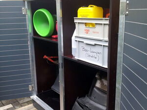 . Ein offener Gartenschrank mit verschiedenem Inhalt: Eimer, Kisten (eine beschriftet mit "Bau-Lampen"), Schneeschieber.