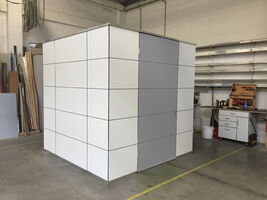 The image shows a small, cubicle-like structure with a grid pattern on its surfaces, located inside a workshop or industrial space. The structure appears to be made of panels, and there is a workbench with tools in the background.