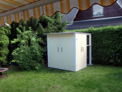 The image shows a small garden shed or storage unit situated on a grassy lawn. It's surrounded by green shrubs or bushes, and there are some trees in the background. Above, there's an awning with a white and yellow striped pattern. The shed has double doors and is positioned near the edge of the yard.