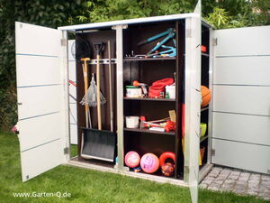 . The image shows a garden storage cabinet with open doors. Inside, you can see various items organized on shelves and racks:

- A snow shovel, rakes, and other garden tools such as a spade and hoe.
- A sports ball (possibly a basketball) and other inflatable balls.
- A bicycle or scooter handlebars are visible on an upper shelf.
- Various garden supplies, possibly including pots, a watering can, and other small tools.
- Containers and storage boxes for organization.

The cabinet is located outdoors on a grassy area with plants and trees in the background.