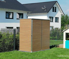 . The image shows a wooden outdoor storage shed placed in a grassy yard, with a residential building in the background. The shed has vertical wooden panels and a simple, modern design. There is also a small white and blue playhouse nearby, and both structures are fenced in.
