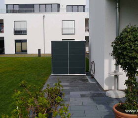 . The image depicts a modern outdoor setting. Elements include:

1. A white multi-story building in the background with balconies and windows.
2. A large green lawn.
3. A paved area with gray tiles.
4. A small tree or shrub in a pot on the right.
5. A dark panel or structure near the wall.
6. Outdoor furniture such as a table and chair.
7. A coiled hose or cable on the wall.