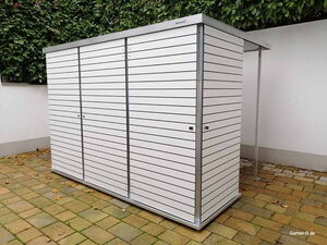 . Ein moderner, weißer Gartenschrank mit Lamellen, steht auf einem gepflasterten Bereich vor grüner Hecke.