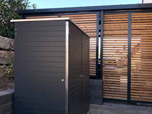 . The image features a small, modern outdoor storage shed or cabinet with a sleek design. It appears to be made of metal or composite materials with horizontal paneling. The setting includes a paved area with stone tiles and a wooden privacy screen fence in the background. There is also some landscaping with plants and mulch nearby.