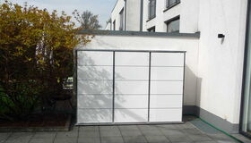 . The image shows a modern outdoor storage unit situated on a tiled patio. It is placed against a wall and is surrounded by some shrubs or small trees on one side. The building is contemporary, with large windows and clean white walls.
