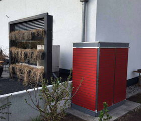 . The image shows a garden scene with a red storage unit on the right side. There is a vertical structure on the left with several levels for planting, but the plants appear dried or withered. In the foreground, there are some small plants or bushes, and the setting is next to a white wall with a drain pipe.