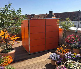 . The image shows a balcony garden with various elements:

1. A bright orange storage unit.
2. Several potted plants and flowers.
3. An orange folding chair.
4. A wooden deck floor.
5. A view of surrounding buildings in the background.

The setting appears vibrant and colorful, designed for an urban outdoor space.