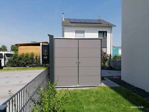 . Ein grauer Lagerschrank in einem Garten, mit Häusern und Solarpanelen im Hintergrund.