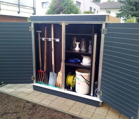 . The image shows an outdoor storage cabinet with its doors open. Inside, there are:

1. Several long-handled garden tools, such as a shovel, rake, fork, and broom.
2. Shelves holding various items, including:
   - Glass vases or pitchers
   - Plates
   - Coiled rope or hose
   - A white bucket containing garden tools
   - Yellow garden hose

The cabinet appears to be neatly organized, with the tools hanging on hooks.