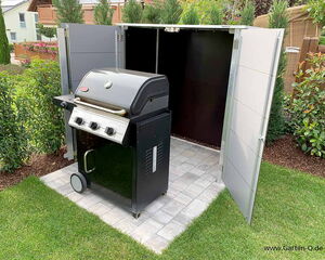 . The image shows a black grill placed on a paved surface with a surrounding enclosure. The enclosure has open doors, revealing the grill inside. The area is surrounded by a well-maintained garden with grass and various bushes.

Here's a breakdown in array format:

- Grill
  - Color: Black
  - Components: Cooking area, knobs, wheels  
 
- Enclosure
  - Doors: Open
  - Material: Metal or similar hard material

- Surroundings
  - Surface: Paved
  - Vegetation: Grass, bushes, and other plants

- Background
  - Fencing: Wooden
  - Additional: Nearby buildings and garden features
