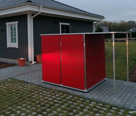 . Ein modernes rotes Gartenhaus neben einem grau gestrichenen Haus mit zwei Fenstern und zwei Töpfen.