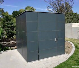 . The image shows a modern, sleek storage shed or garden structure with a flat roof and smooth, dark panels. It's located in a landscaped garden with paved pathways and surrounded by greenery and trees.