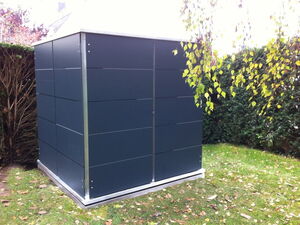 . The image shows a modern-looking shed in a garden. It's constructed with dark panels and has a metallic frame. The shed is surrounded by grass and trees, and there are some falling leaves on the ground, indicating it might be autumn.