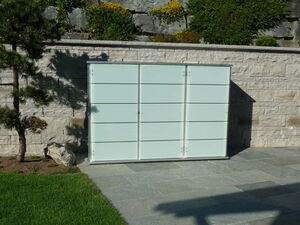 . The image shows a modern outdoor setting with a sleek storage unit or cabinet against a stone wall. The unit has frosted glass panels, and the area is landscaped with a tree and some greenery. The ground is covered with flat stone tiles, and there is a grassy area nearby.