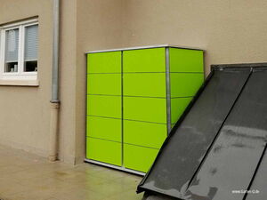 . The image shows a bright green storage cabinet or shed placed against a beige wall. There's a gray downspout on the left side of the image, next to a window with closed blinds. Nearby, there's a sloped black surface, possibly a part of a roof.