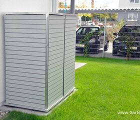 . The image features an outdoor storage unit with a sleek, modern design, positioned next to a building. The unit is made of horizontal panels, possibly metal or plastic. In the background, there are two parked cars and a fence, with grass in the foreground. The setting appears to be a residential area.
