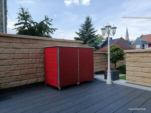 . The image shows a red outdoor storage unit or cabinet placed on a patio. It’s positioned against a brick wall. There’s a lamp post with three lamps next to it, and a small tree in a planter. The background features another brick wall, some bushes, and a glimpse of nearby buildings.