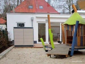 . Ein moderner Gartenschrank und ein Holzspielhaus auf einem Sandspielplatz vor einem weißen Gebäude.