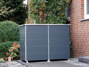 . The image shows a sleek, modern garden storage unit positioned next to a brick wall. It’s a dark grey rectangular structure with a metal frame and a flat top. The unit is situated in a garden area, surrounded by bushes and plants.