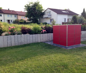 . Ein Garten mit einem grauen Zaun, blühenden Sträuchern und einem roten quadratischen Schuppen im Vordergrund.