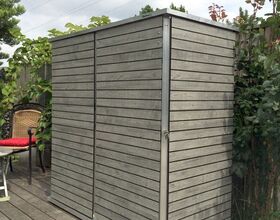 . The image shows a small, rectangular storage shed made of horizontal wooden planks with a metal frame. It's positioned on a wooden deck with some outdoor plants and garden furniture visible nearby. The surroundings include a fence and greenery, and it's set against a partly cloudy sky.