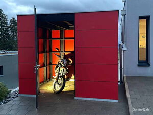 . Ein Motorrad steht in einem beleuchteten, offenen roten Schuppen oder Unterstand.
