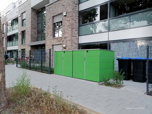 . In the image, there's a modern-looking apartment building with large windows and a brick facade. In front of the building, there is a green structure, possibly a storage unit or bin enclosure. Next to it are three black bins with blue lids. A paved walkway runs in front of these structures, and there is a small fenced area with some bicycles. A patch of greenery with a tree is in the foreground.
