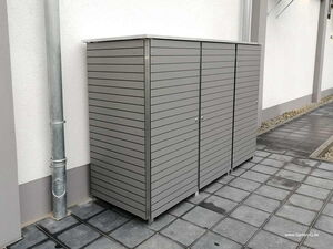 . Ein grauer Gartenschrank aus Metall steht auf gepflastertem Boden an einer weißen Wand.