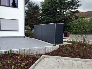 . Moderner Hinterhof mit grauem Schuppen, Pflastersteinen, Gartenbeeten und Bäumen neben einem Haus.