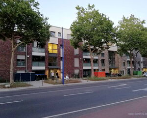 The image shows a residential building with several apartments and a row of trees along the sidewalk. The building has a modern design with large windows and brickwork. There are people walking near the building, and bicycles can be seen parked nearby. The street in front of the building is relatively wide, with markings and lanes visible.