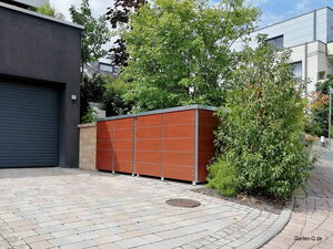 . Ein moderner Gartenschuppen neben einer Garage, umgeben von Pflanzen und Pflastersteinen.